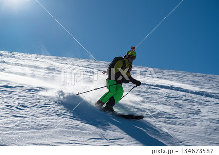 Skier skiing off piste through fresh powder on a sunny mountain slope, kicking up snow as she descends, creating a dynamic and energetic winter sports action scene under a clear blue sky Skier skiing off piste through fresh powder on a sunny mountain slope, kicking up snow as she descends, creating a dynamic and energetic winter sports action scene under a clear blue sky 134678587