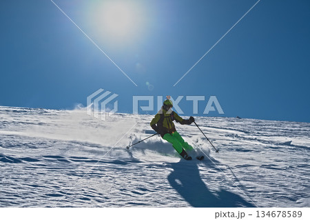 Skier skiing off piste through fresh powder on a sunny mountain slope, kicking up snow as she descends, creating a dynamic and energetic winter sports action scene under a clear blue sky 134678589