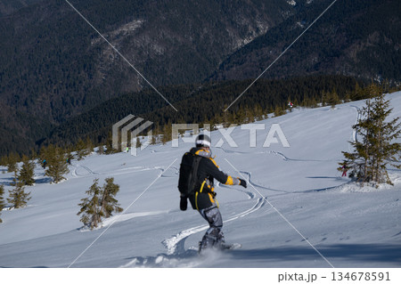 A snowboarder in yellow and grey camouflage gear rides down a pristine snowy slope on a sunny winter day, with a scenic mountain forest background 134678591