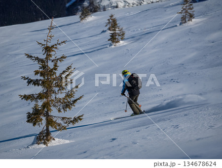Skier skiing off piste through fresh powder on a sunny mountain slope, kicking up snow as she descends, creating a dynamic and energetic winter sports action scene under a clear blue sky 134678624