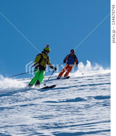 Two male skiers descending a snowy slope in unison under a clear blue sky, kicking up powder snow, with distant pine trees on the horizon 134678748