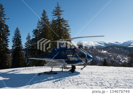A blue helicopter awaits on a snowy mountain, ready for heli-skiing. Surrounded by pine trees and stunning alpine scenery under a clear blue sky, it captures the essence of winter adventure A blue helicopter awaits on a snowy mountain, ready for heli-skiing. Surrounded by pine trees and stunning alpine scenery under a clear blue sky, it captures the essence of winter adventure 134678749