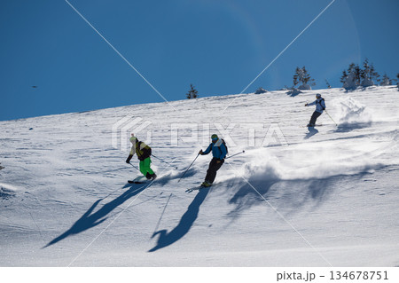 Three skiers carve down a sunny, powder-covered mountain slope, kicking up fresh snow under a clear blue sky, creating a dynamic winter sports action scene Three skiers carve down a sunny, powder-covered mountain slope, kicking up fresh snow under a clear blue sky, creating a dynamic winter sports action scene 134678751