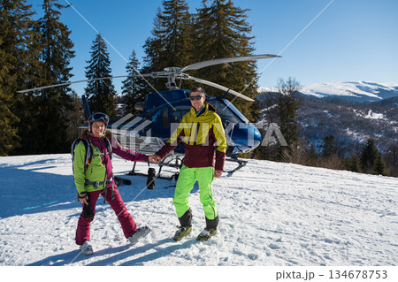 A smiling man and woman in bright ski gear stand on snowy terrain beside a blue helicopter, celebrating a heliskiing adventure in sunny alpine mountains 134678753