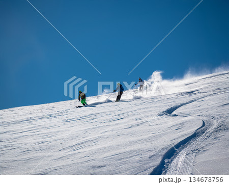 Three skiers carve down a sunny, powder-covered mountain slope, kicking up fresh snow under a clear blue sky, creating a dynamic winter sports action scene 134678756