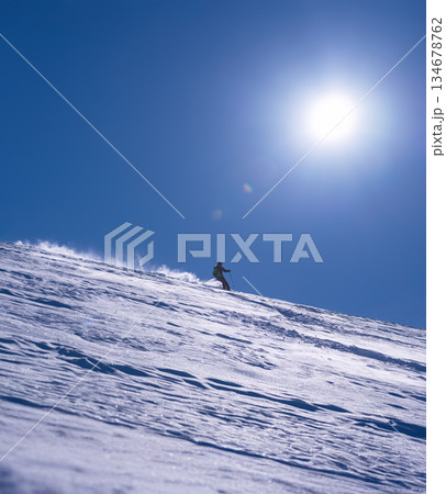 A female skier carves through fresh powder on a sunny mountain slope, kicking up snow as she descends, creating a dynamic and energetic winter sports action scene under a clear blue sky 134678762