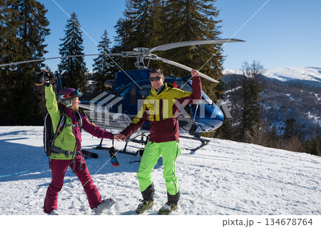 A smiling man and woman in bright ski gear stand on snowy terrain beside a blue helicopter, celebrating a heliskiing adventure in sunny alpine mountains 134678764