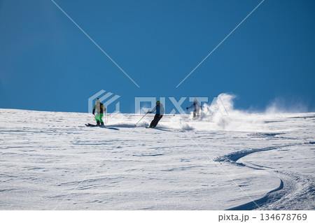 Three skiers carve down a sunny, powder-covered mountain slope, kicking up fresh snow under a clear blue sky, creating a dynamic winter sports action scene 134678769