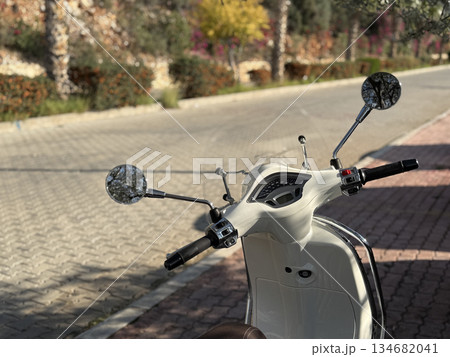 Cream-colored scooter parked on sunlit cobblestone street with blurred trees in background Cream-colored scooter parked on sunlit cobblestone street with blurred trees in background 134682041