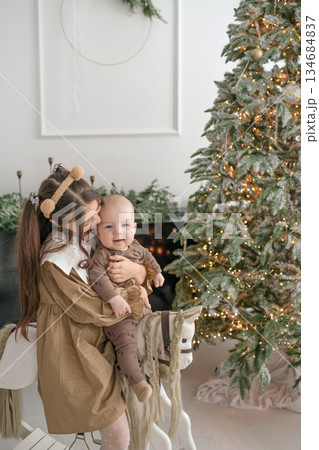 A girl holds her little brother in arms, swinging on a toy wooden horse. New Year's Holiday tree behind the children A girl holds her little brother in arms, swinging on a toy wooden horse. New Year's Holiday tree behind the children 134684837