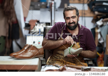 Smiling shoemaker at work in leather workshop measuring a shoe with a tape, showcasing handmade craftsmanship Smiling shoemaker at work in leather workshop measuring a shoe with a tape, showcasing handmade craftsmanship 134686180