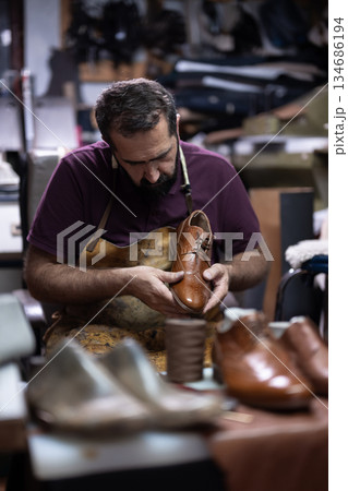 Cobbler expertly crafts a brown leather shoe in a bustling workshop environment 134686194