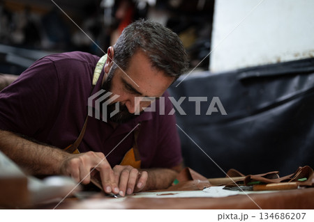 A skilled craftsman with a beard focuses on leather cutting at a workshop A skilled craftsman with a beard focuses on leather cutting at a workshop 134686207