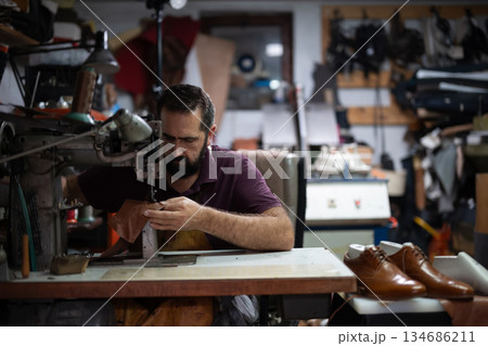 Cobbler sewing leather in a busy workshop with leather shoes on the table nearby Cobbler sewing leather in a busy workshop with leather shoes on the table nearby 134686211