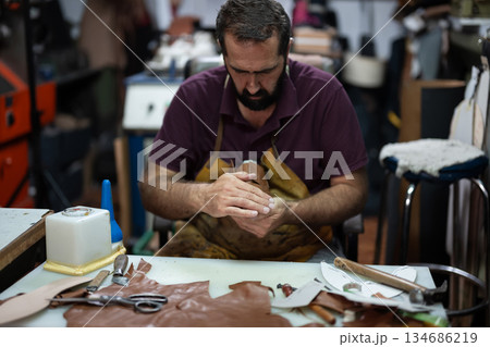 Leather artisan shapes leather at a cluttered workshop bench, focused on handcrafting a piece 134686219