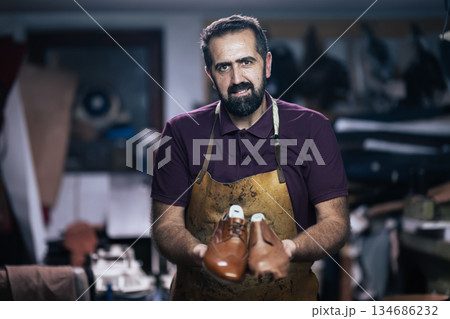 Skilled craftsman in a workshop presenting brown leather shoes, wearing an apron and focused on his craft Skilled craftsman in a workshop presenting brown leather shoes, wearing an apron and focused on his craft 134686232