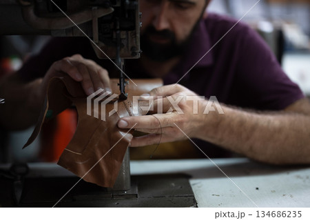 Leather work in progress: skilled craftsman sewing brown leather piece with industrial sewing machine Leather work in progress: skilled craftsman sewing brown leather piece with industrial sewing machine 134686235