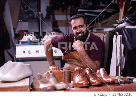 Shoemaker in a leather workshop, smiling at his bench with polished brown shoes 134686236