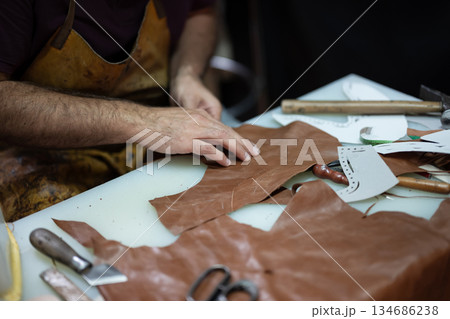 Leather craftsman at work shaping brown leather pieces with tools and patterns on a busy worktable 134686238