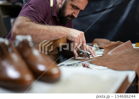 Skilled leather craftsman cutting leather pieces at a worktable in a busy workshop. 134686244