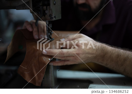 Craftsman guides brown leather under a sewing machine, stitching a piece with careful, precise handiwork. 134686252