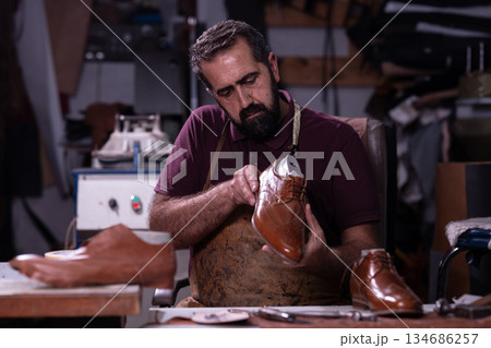 Cobbler crafts brown leather shoes in a workshop, focusing on detailing and shaping Cobbler crafts brown leather shoes in a workshop, focusing on detailing and shaping 134686257