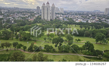 A glowing golf course with sand bunkers and floodlights stretches beneath ornate towers and a vivid sunset, blending sport, architecture, and nature in a dramatic dusk cityscape in Jakarta A glowing golf course with sand bunkers and floodlights stretches beneath ornate towers and a vivid sunset, blending sport, architecture, and nature in a dramatic dusk cityscape in Jakarta 134686267