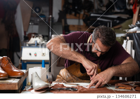 Man crafts leather shoes in a cluttered workshop, focused on stitching and detailing 134686271