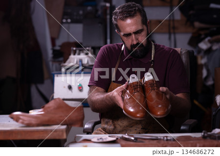 Shoemaker in a workshop holds polished brown leather shoes, preparing them for repair Shoemaker in a workshop holds polished brown leather shoes, preparing them for repair 134686272