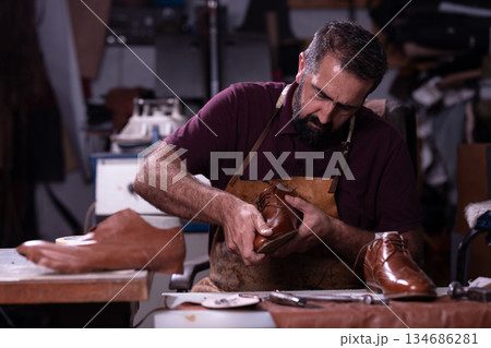 Leather craftsman shaping a brown leather shoe in a busy workshop with tools and boots nearby Leather craftsman shaping a brown leather shoe in a busy workshop with tools and boots nearby 134686281
