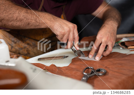 Leather craft in progress: close-up of hands cutting leather with a knife and tools Leather craft in progress: close-up of hands cutting leather with a knife and tools 134686285