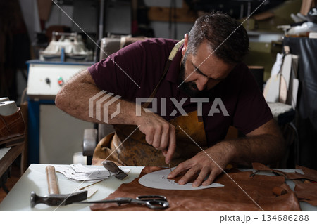 Skilled craftsman carving leather at a worktable with tools and leather scraps nearby Skilled craftsman carving leather at a worktable with tools and leather scraps nearby 134686288