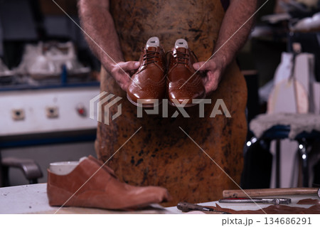 Cobbler holds polished brown leather dress shoes in workshop with tools on the workbench 134686291