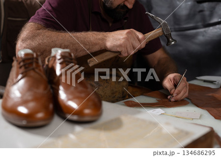 Cobbler at work shaping leather with hammer and chisel, crafting a pair of brown shoes 134686295