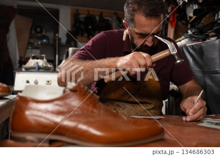 Cobbler at work repairing leather shoe with hammer and awl in a focused old-world workshop Cobbler at work repairing leather shoe with hammer and awl in a focused old-world workshop 134686303