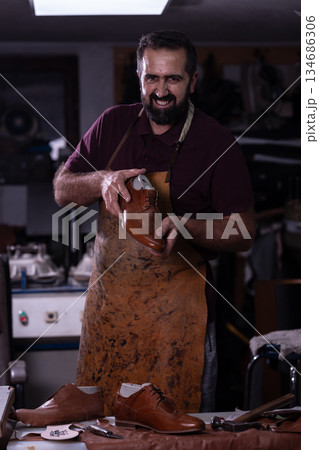 Cobbler in a leather apron polishing a brown shoe in his busy workshop 134686306