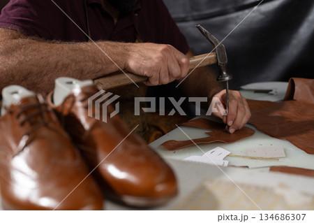 Leather craftsman hammering leather to shape pieces for shoes on the bench, with polished brown shoes nearby Leather craftsman hammering leather to shape pieces for shoes on the bench, with polished brown shoes nearby 134686307
