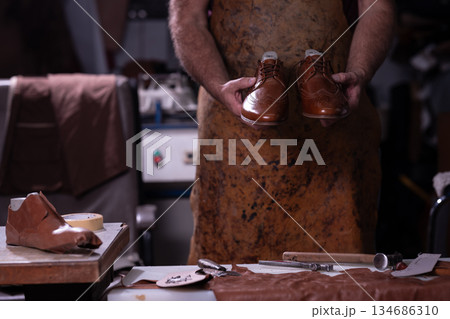 Craftsman holds polished brown leather shoes in a busy workshop, surrounded by tools and leather scraps 134686310