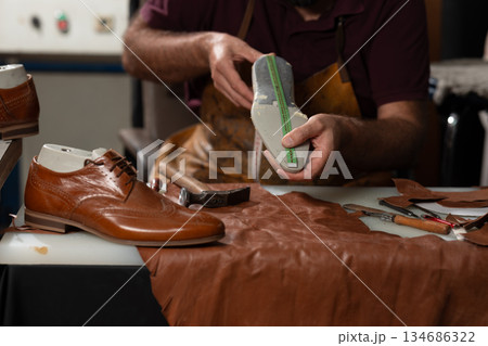 Skilled artisan examines a leather sole with a measuring tape in a busy shoemaking workshop 134686322