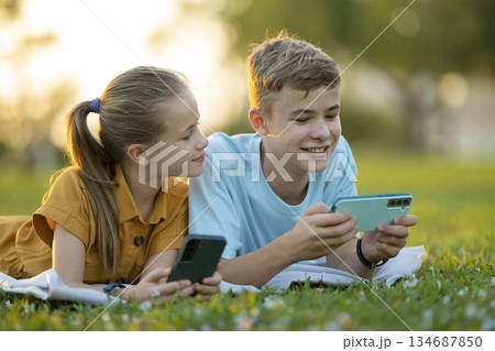 Positive children boy and girl playing online games on their mobile phones with friends outdoors in summer park. Positive children boy and girl playing online games on their mobile phones with friends outdoors in summer park. 134687850