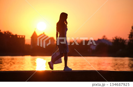 Lonely young woman walking alone on lake shore enjoying warm evening. Wellbeing and relaxing in nature concept. 134687925