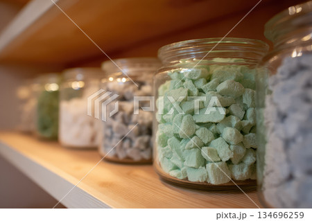 Row of clear glass jar filled with green blue white colorful biodegradable material rock placed wooden shelf inventory eco friendly shop displaying organized clean minimalist store atmosphere 134696259