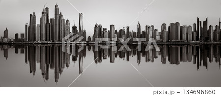 Monochrome city skyline with tall skyscrapers reflecting on calm water under a cloudy sky, Dubai Marina bay UAE 134696860