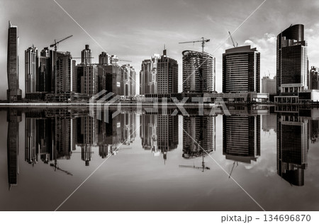 Monochrome city skyline with tall skyscrapers reflecting on calm water under a cloudy sky, Dubai Marina bay UAE 134696870