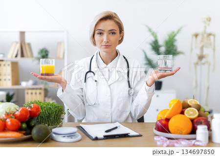 A female doctor holds a glass of juice and water, surrounded by fruits and vegetables, promoting healthy choices. A female doctor holds a glass of juice and water, surrounded by fruits and vegetables, promoting healthy choices. 134697850