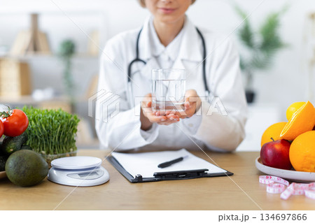 A doctor holding a glass of water, surrounded by fresh fruits and vegetables, promoting a healthy lifestyle. A doctor holding a glass of water, surrounded by fresh fruits and vegetables, promoting a healthy lifestyle. 134697866