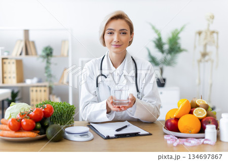 A nutritionist holds a glass of water, surrounded by fresh fruits and vegetables, promoting healthy eating habits. A nutritionist holds a glass of water, surrounded by fresh fruits and vegetables, promoting healthy eating habits. 134697867