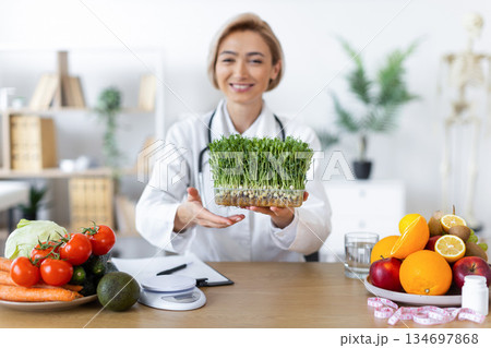 A smiling nutritionist presents a tray of microgreens, surrounded by fresh fruits and vegetables, promoting healthy eating. 134697868