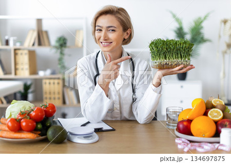 A smiling nutritionist points to a tray of microgreens, surrounded by fresh fruits and vegetables, promoting healthy eating. 134697875