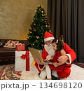 Santa Claus sitting with young child on lap while using laptop computer near festive decorated Christmas tree and wrapped presents in living room at home 134698120